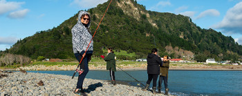 Four people are fishing with rods on a rocky shore. A green hill is in the background.