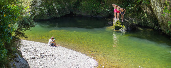 Kids jumping off a rock into a river and a person sitting on the shore watching