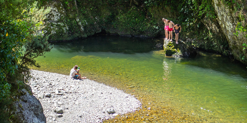 Kids jumping off a rock into a river and a person sitting on the shore watching