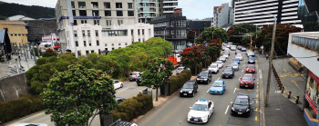 Photo of many cars stuck in slow moving traffic, in central Wellington. There are many tall buildings surrounding the busy six lane road, and some trees adjacent to the road. 