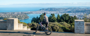 Photo of a mountain biker at a lookout, overlooking the city and harbour of Dunedin. It is a sunny, blue sky day. 