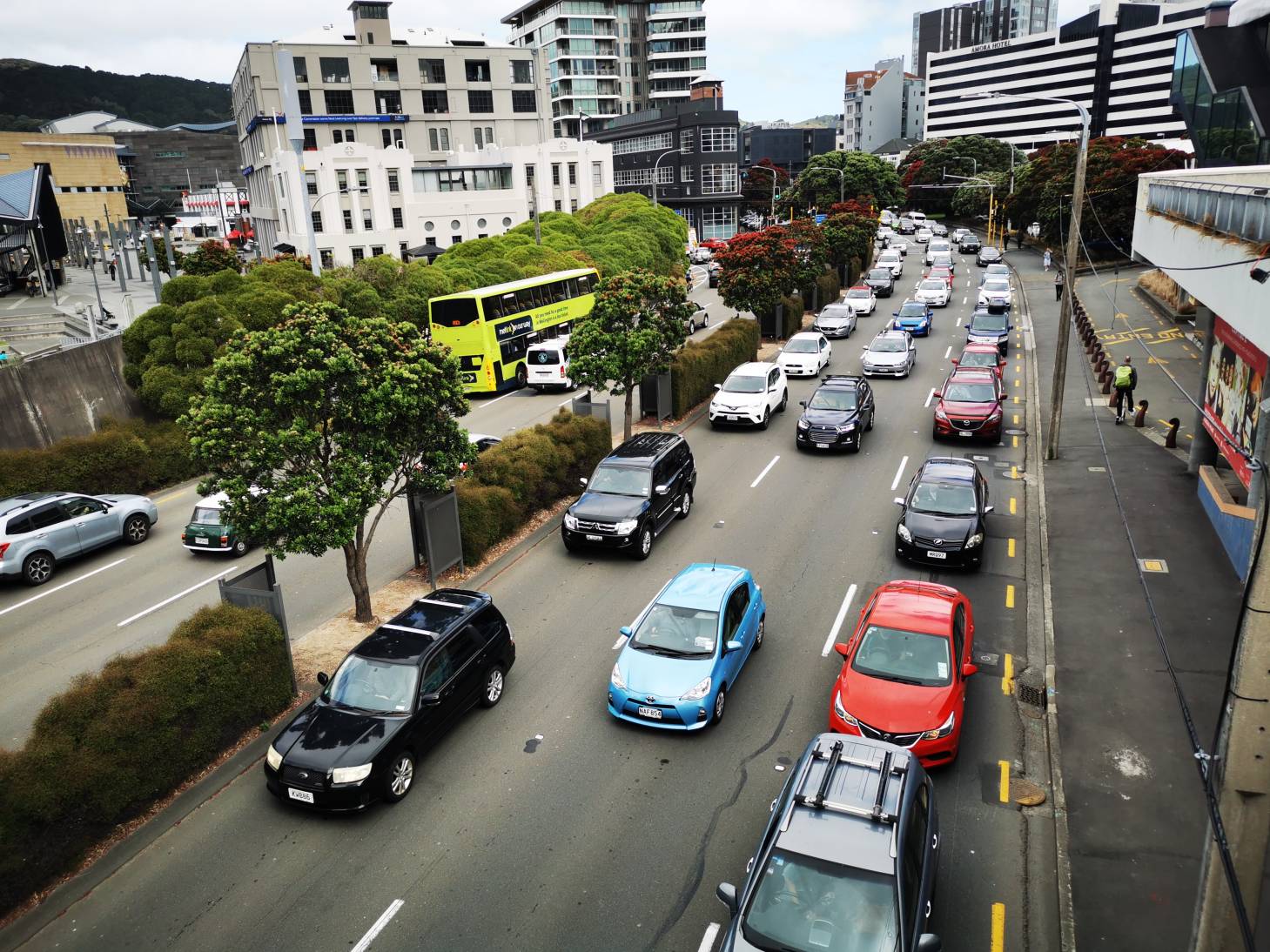 Photo of many cars stuck in slow moving traffic, in central Wellington. There are many tall buildings surrounding the busy six lane road, and some trees adjacent to the road.