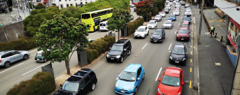 Photo of many cars stuck in slow moving traffic, in central Wellington. There are many tall buildings surrounding the busy six lane road, and some trees adjacent to the road.