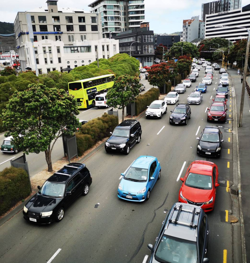 Photo of many cars stuck in slow moving traffic, in central Wellington. There are many tall buildings surrounding the busy six lane road, and some trees adjacent to the road.