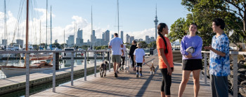 In the foreground, three young adults stand on a walkway, chatting. In the mid-ground, a family are walking their dogs. In the background are trees, Auckland's sky tower, buildings and the viaduct.
