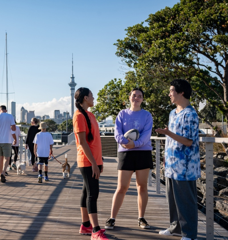 In the foreground, three young adults stand on a walkway, chatting. In the mid-ground, a family are walking their dogs. In the background are trees, Auckland's sky tower, buildings and the viaduct.