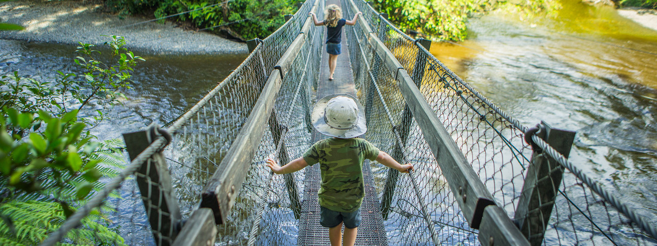 Two kids running along a suspension bridge over a river and surrounded by plants and trees.