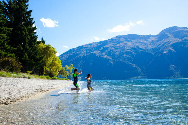 Two kids in wetsuits running into the water of a serene lake on a sunny day.