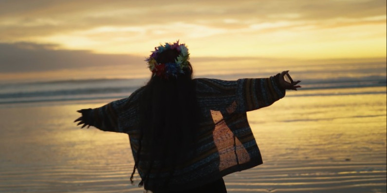 A Pasifika person standing in front of the ocean with their arms open wide.