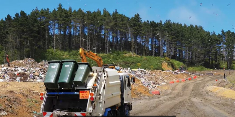 Landscape of a landfill, with a truck driving past with bins of rubbish on the back.