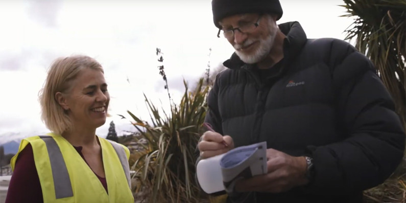 A woman in hi-vis talking to a man in a puffer jacket who is writing down information.