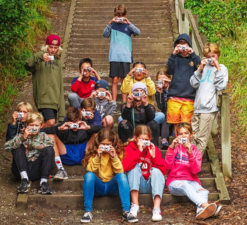 A group of kids sitting on some steps, all holding up cameras as if they are taking photos.